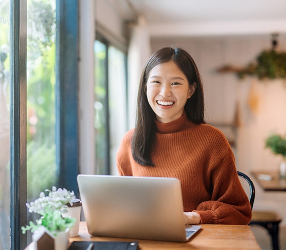A young female wearing an orange sweater, sitting behind a laptop, and smiling after seeing a Delta Dental dentist in Fort Worth